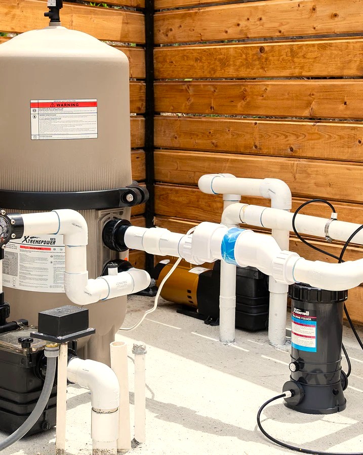 Pool filtration system with a large tan sand filter, white PVC pipes, and a black pump on a concrete pad against a wooden fence.